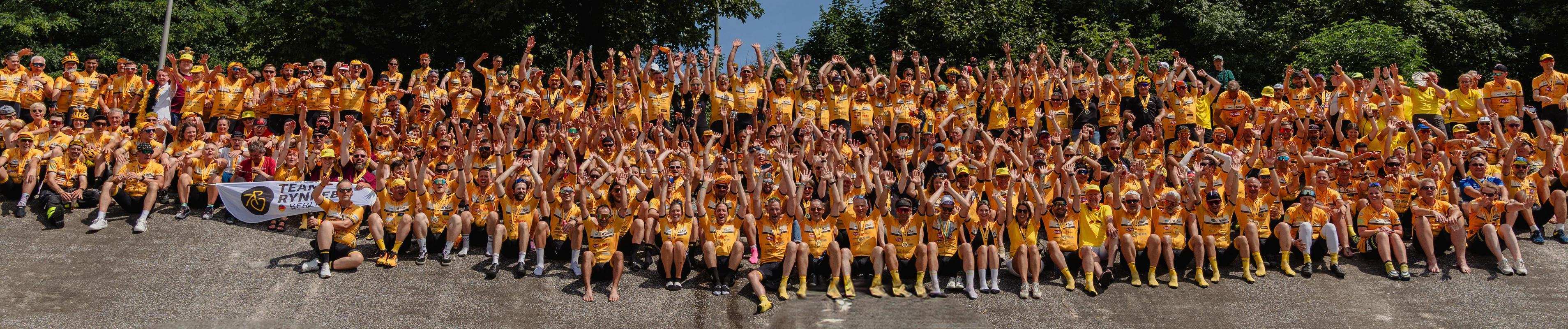 Large group of people in yellow shirts, sitting with hands raised, on a concrete slope with trees in the background.