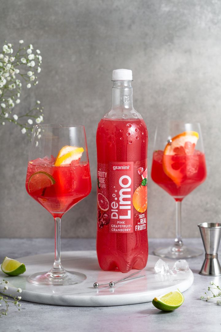A bottle of pink grapefruit cranberry drink on a marble tray, surrounded by two glasses filled with ice, citrus slices, and flowers.