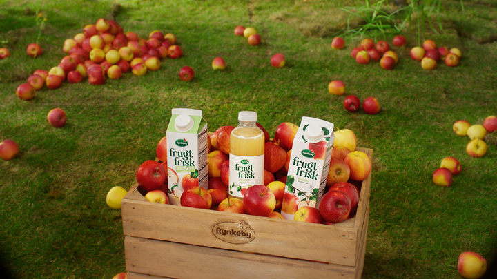 A wooden crate filled with apples and Rynkeby Frugt & Frisk apple juice containers, placed on grassy ground surrounded by scattered apples.