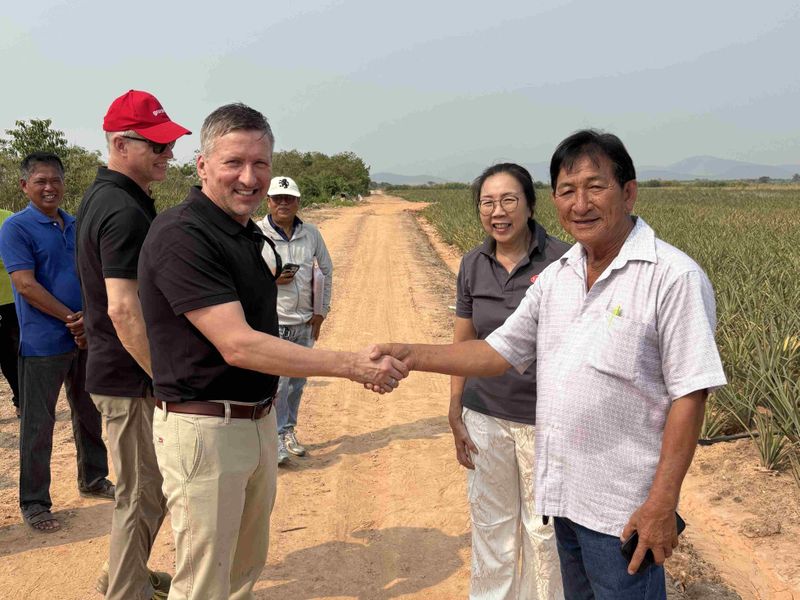 Two men shake hands on a dirt road in a rural area, surrounded by three others. They stand near a field, with mountains in the distance.