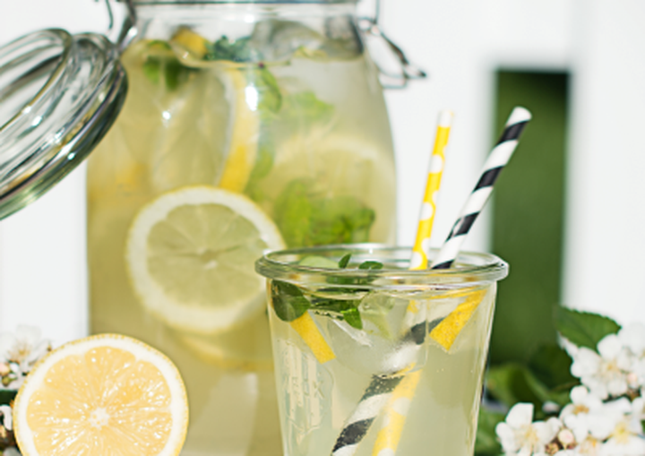 A jar and glass of lemonade with lemon slices, mint leaves, and striped straws, surrounded by flowers on a sunny day.