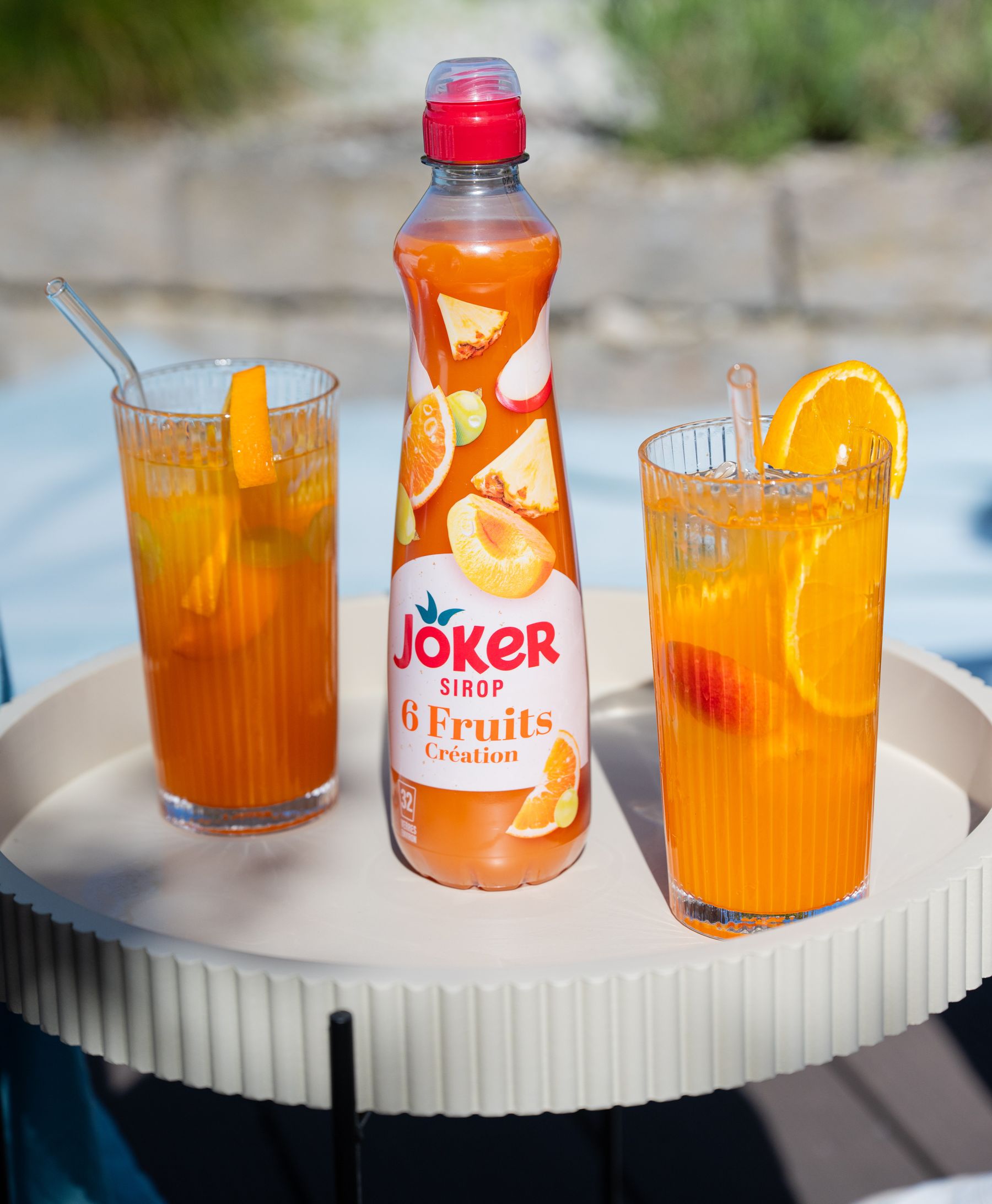 Two glasses of orange drink with slices of orange and a bottle of Joker 6 Fruits syrup on a round tray, outdoors on a sunny day.