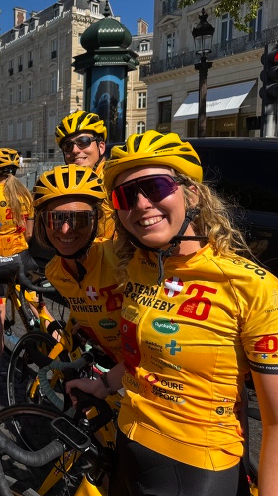 Three smiling cyclists in yellow jerseys and helmets stand beside their bikes on a city street, with historic buildings in the background.