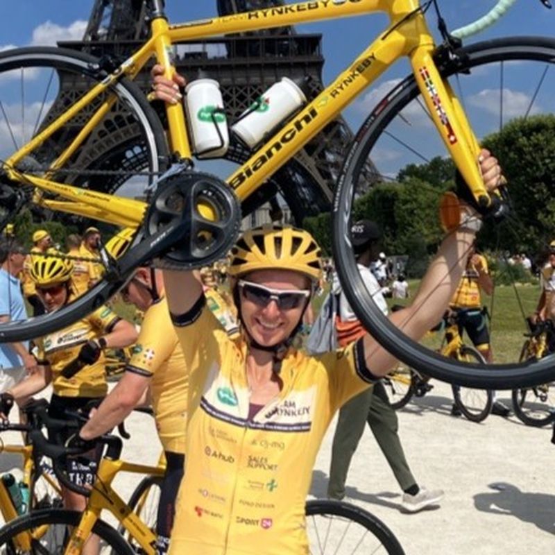 Cyclist in yellow jersey and helmet holding up a bike triumphantly near the Eiffel Tower, with other cyclists and spectators nearby.