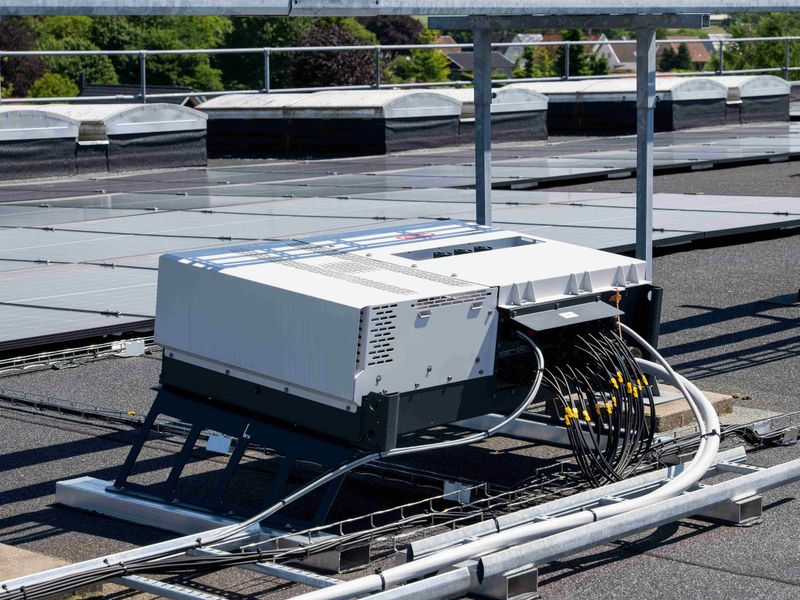 A rooftop solar panel installation with connected wiring and cables on a sunny day, surrounded by greenery in the background.
