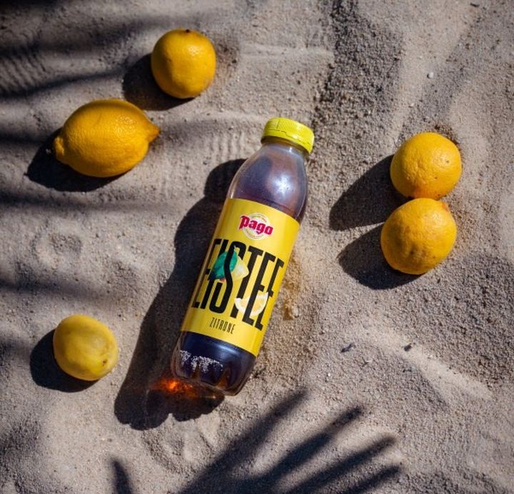 Bottle of iced tea on sandy ground surrounded by lemons, with a shadow of a hand reaching towards it.