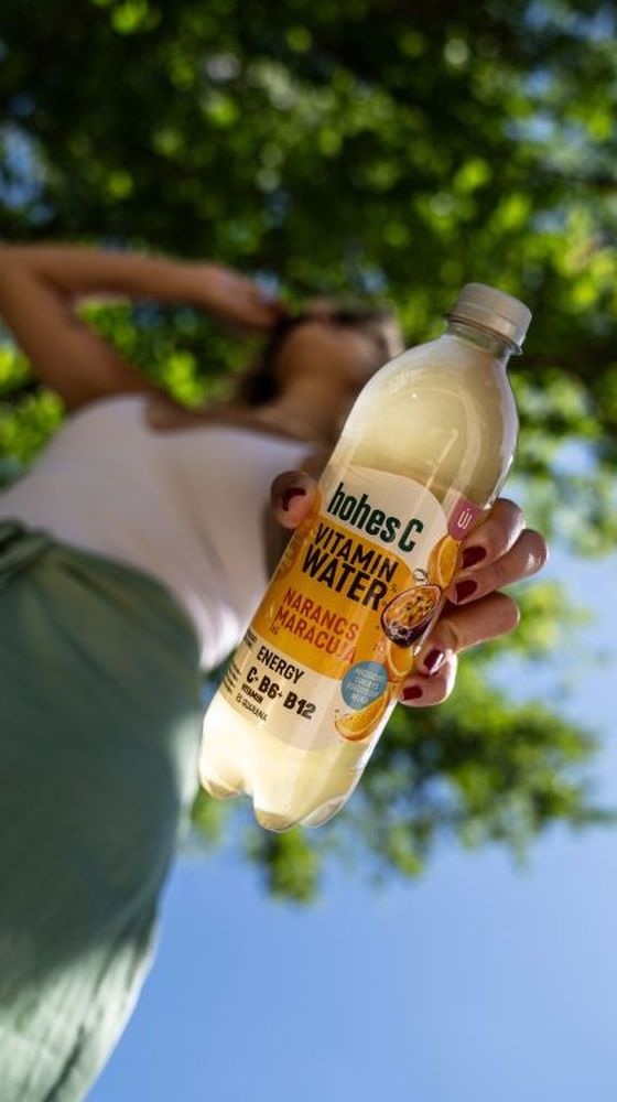 Low-angle view of a person holding a bottle of "hohes C" vitamin water with orange and passion fruit flavor, with trees in the background.