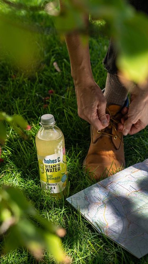 Person tying brown boots next to a map and a bottle of vitamin water on green grass, partially framed by leaves.