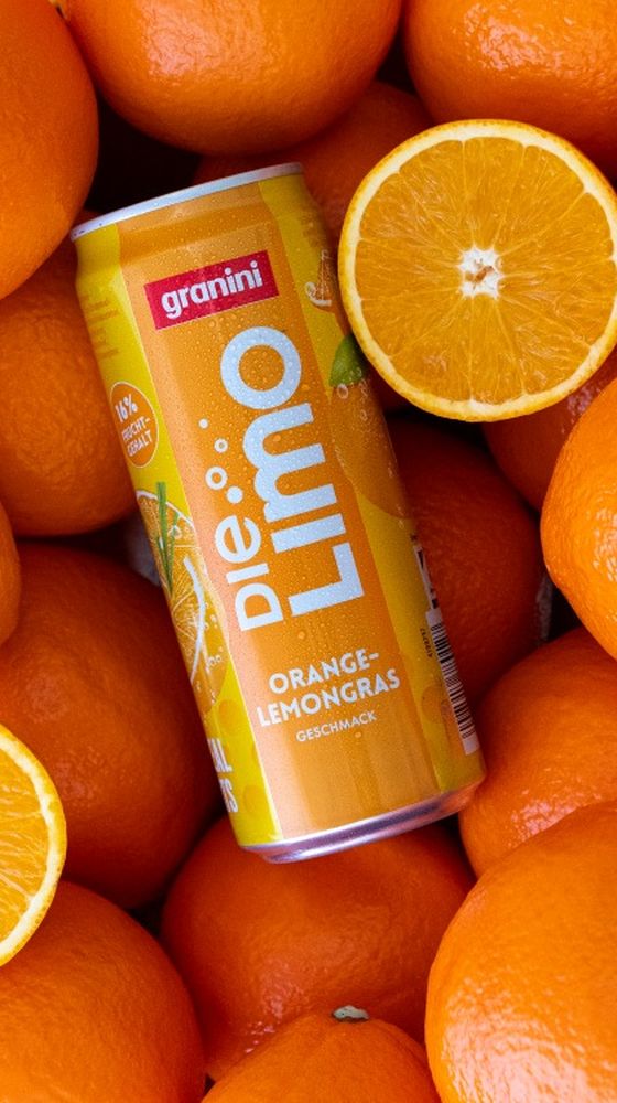 A can of "Die Limo" orange-lemongrass drink surrounded by fresh oranges and two halved oranges, showcasing vibrant, juicy textures.