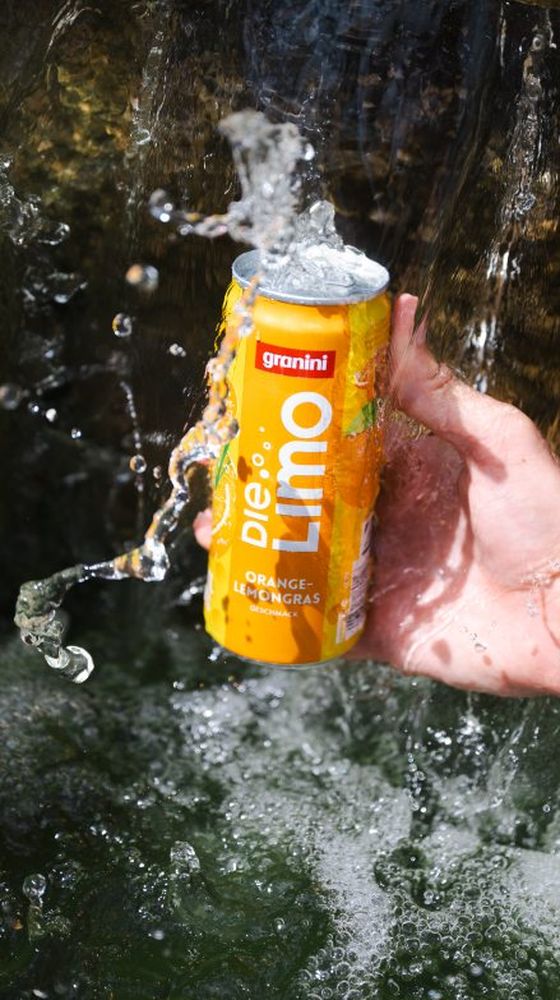 Hand holding a can of "granini Die Limo" orange-lemongrass flavor, splashed by water in a refreshing scene.