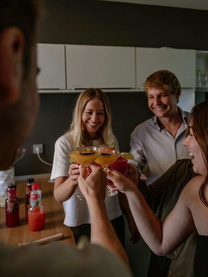 Four people clinking glasses with cocktails in a kitchen, smiling and celebrating. Bottles of mixer drinks are visible on the counter.