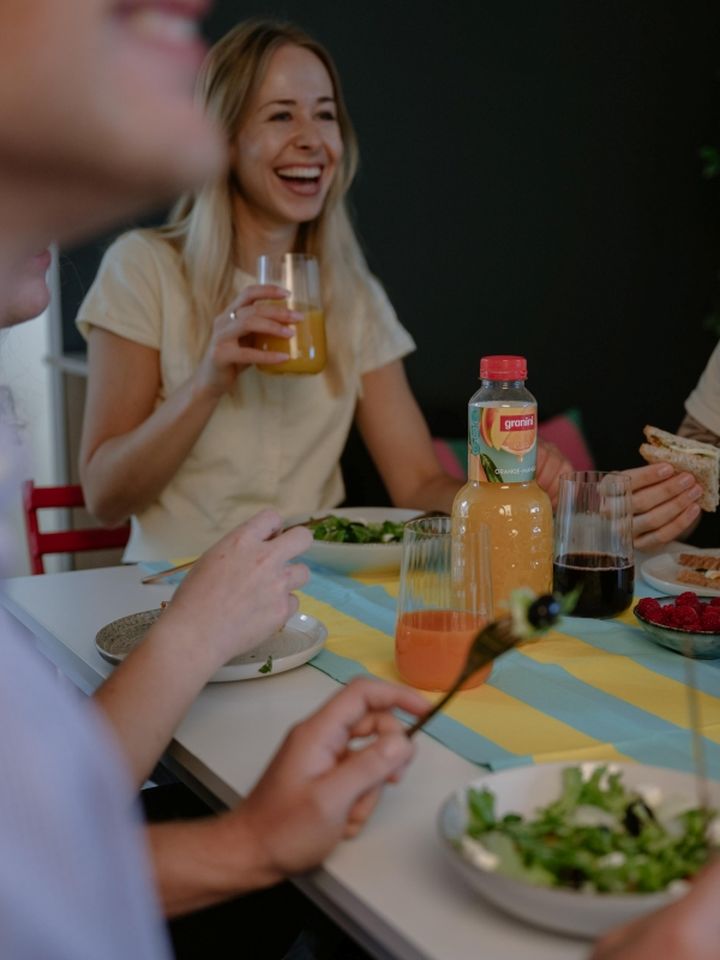 People enjoying a meal at a table with salad, juice, and fruit. A smiling woman holds a glass of orange juice.