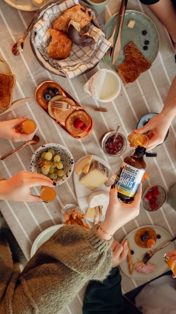 People toasting with orange shots over a breakfast spread featuring pastries, cheese, fruit, and jam on a striped tablecloth.
