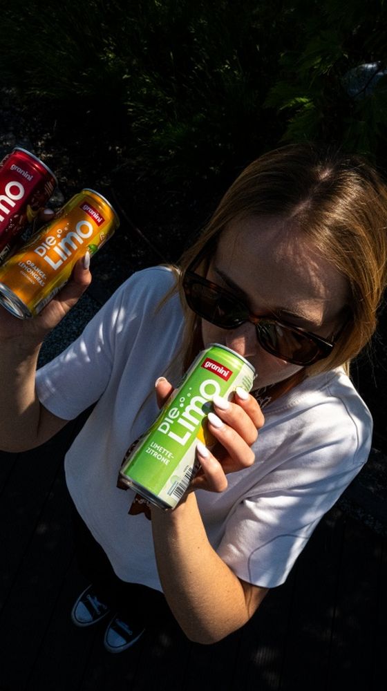 Person in sunglasses drinking a green can of "Die Limo," holding two more cans, one red and one yellow, outdoors.