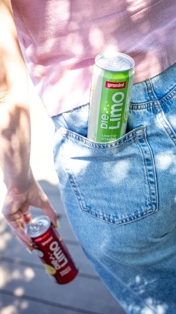 Person in jeans holds a red soda can and has a green one in the back pocket, with dappled sunlight casting shadows.
