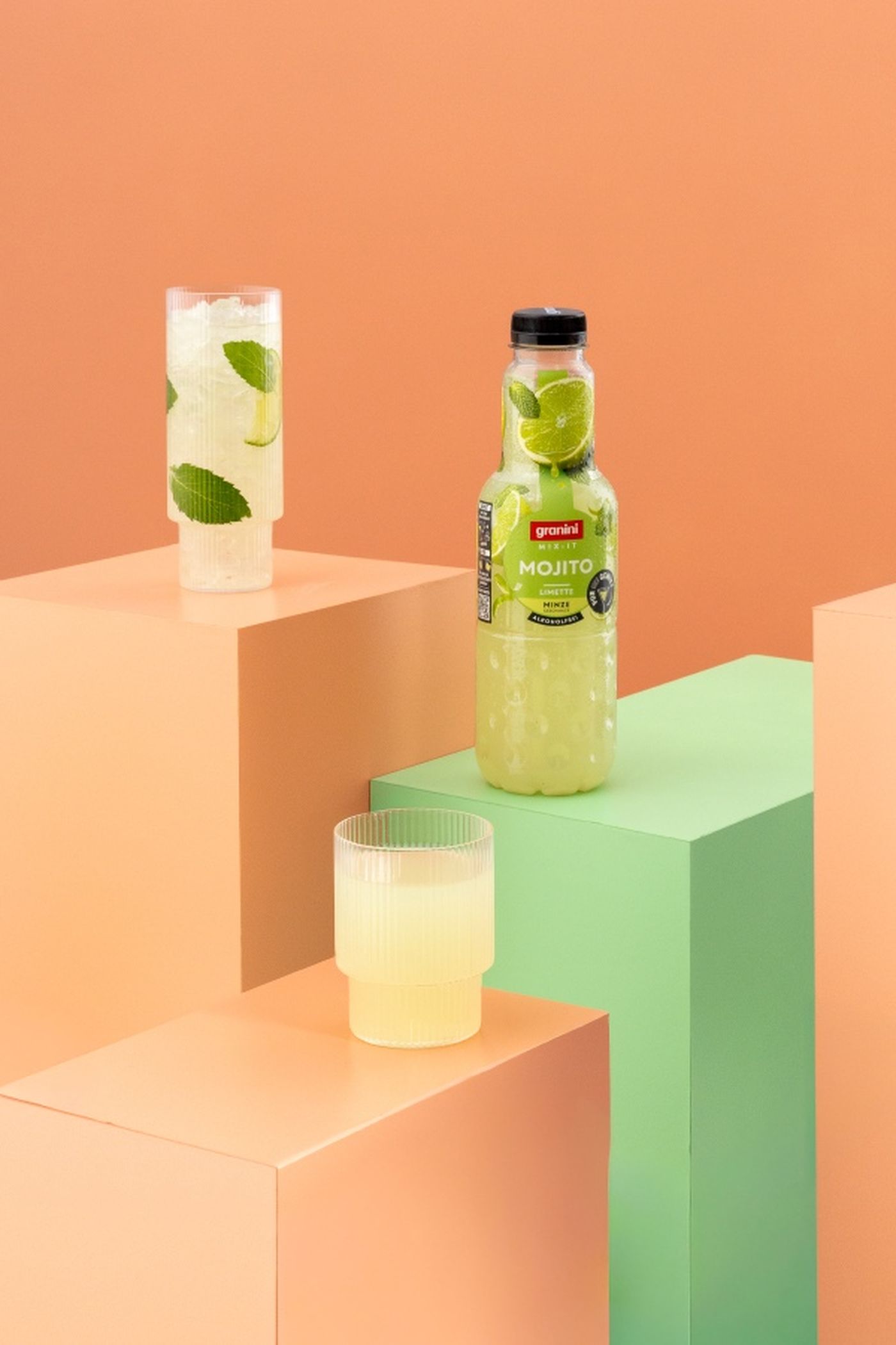 Bottled mojito, two glasses with mint leaf and lime, on pastel geometric blocks against peach background.