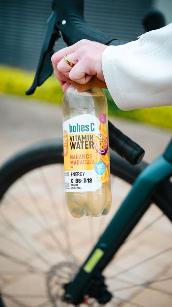 Person holding a bottle of Hohes C Vitamin Water next to a bicycle, featuring orange and maracuja flavors with vitamins C, B6, and B12.