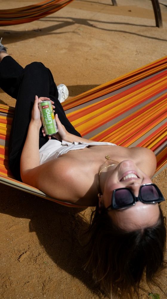 A person relaxes in a colorful hammock, smiling and holding a can of lemon drink, wearing sunglasses and casual clothing.