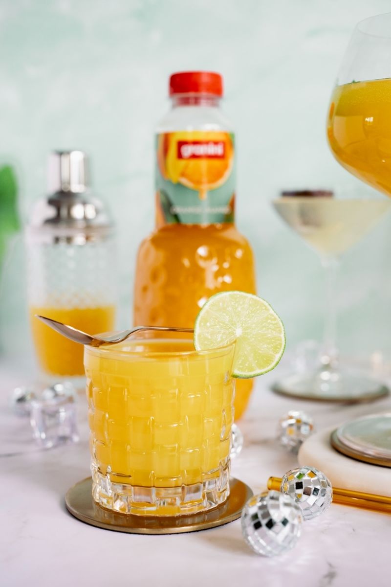 Virgin Margarita with a lime slice, a bottle of juice, cocktail shaker, and disco ball decorations on a marble surface.