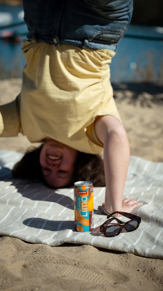 Person doing a headstand on a beach towel with a can of orange drink and sunglasses in front.