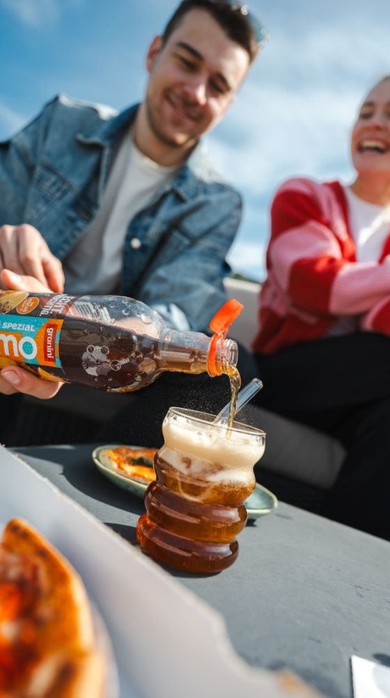 Two people enjoy a sunny day as one pours a fizzy drink into a glass. A plate of pizza is visible nearby.