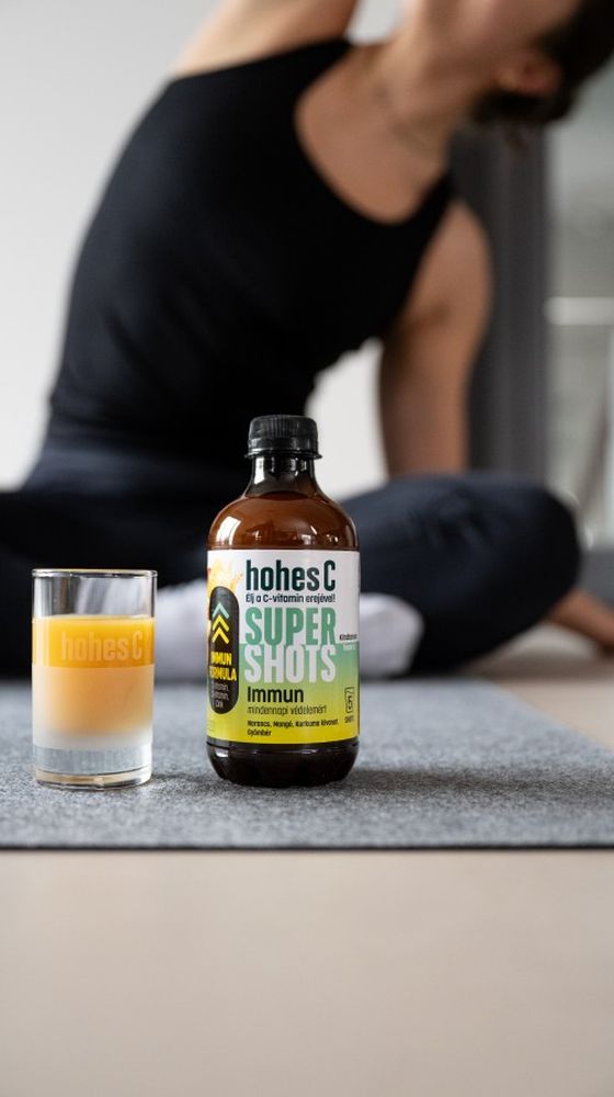 A bottle of hones C Super Shots and a glass sit on a yoga mat, with a person in a black outfit stretching in the background.