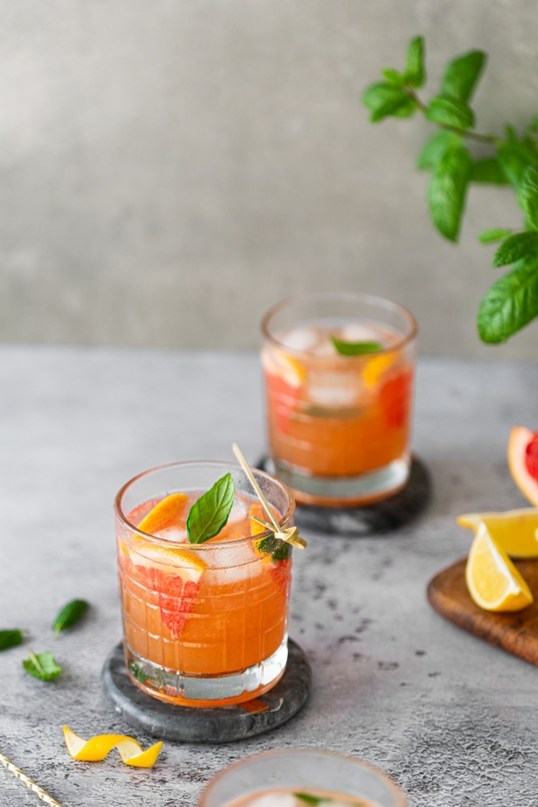 Two glasses of orange and grapefruit cocktails with ice, garnished with mint, on a gray surface; citrus slices nearby.