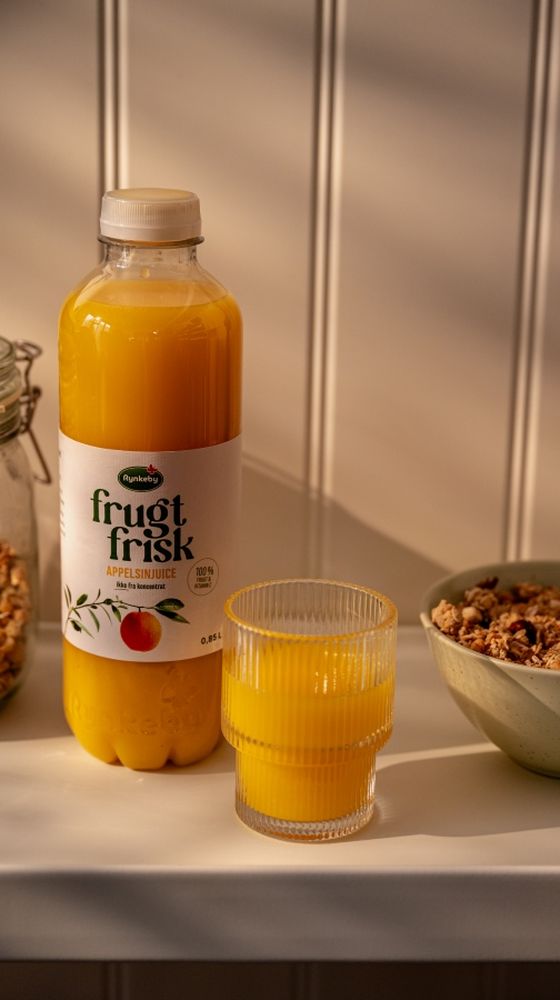 A bottle and glass of orange juice next to a jar of cereal and a bowl of granola on a white surface, with warm light and striped shadows.