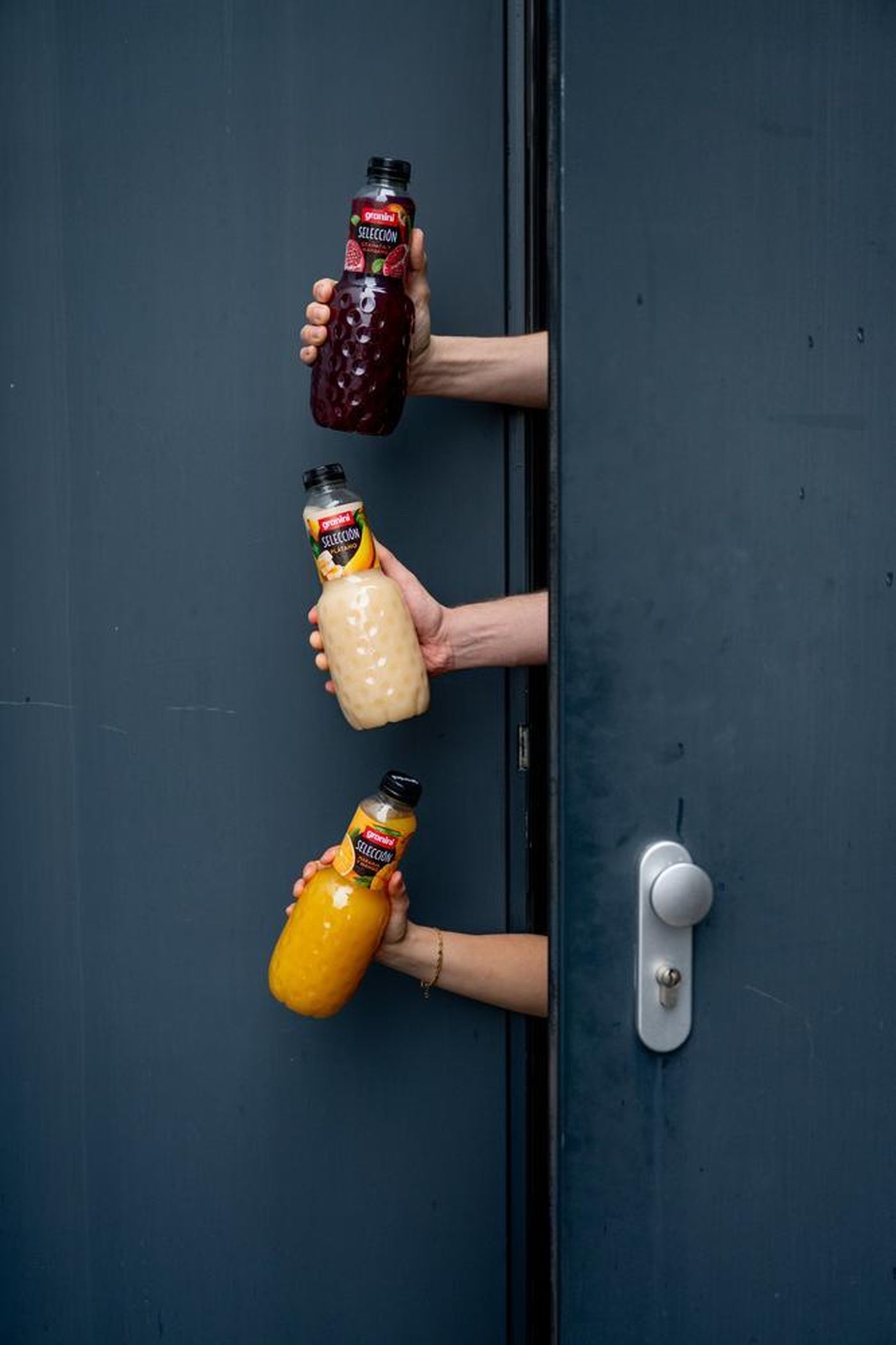 Three arms hold bottles of juice in different colors through a narrow door opening, set against a dark blue background.