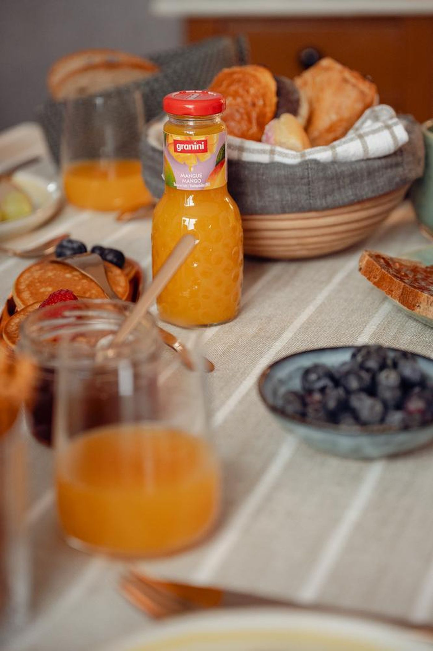 Breakfast table with mango juice, a basket of bread rolls, pancakes topped with berries, jam, and a bowl of blueberries on a striped cloth.