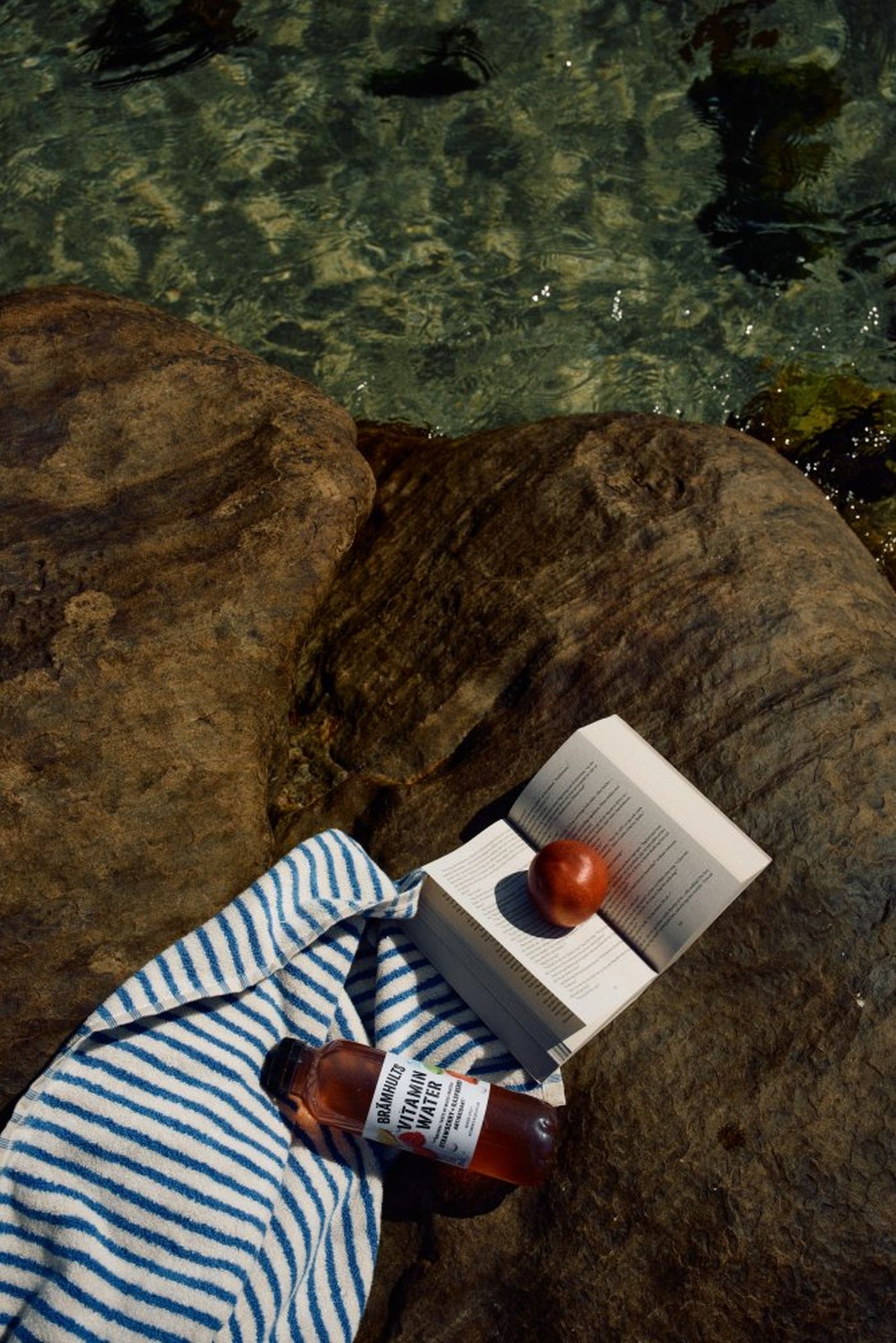 A striped towel, book, nectarine, and vitamin water rest on a rock near clear water, suggesting a relaxing outdoor setting.