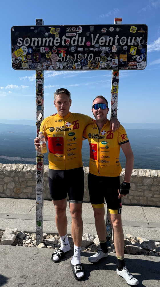 Two cyclists in yellow jerseys stand with arms around each other under a Ventoux summit sign, against a backdrop of a clear sky and distant mountains.