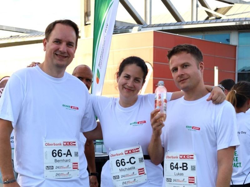 Three people wearing race bibs and white t-shirts pose together, smiling. The person in the middle holds a water bottle.