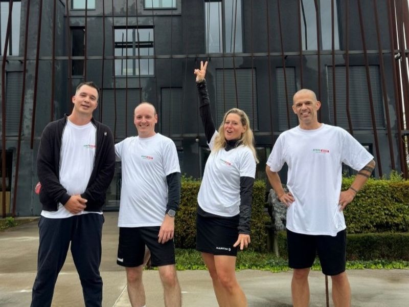 Four people in matching white t-shirts pose cheerfully outside a modern building, with one person raising their arm in a victory sign.