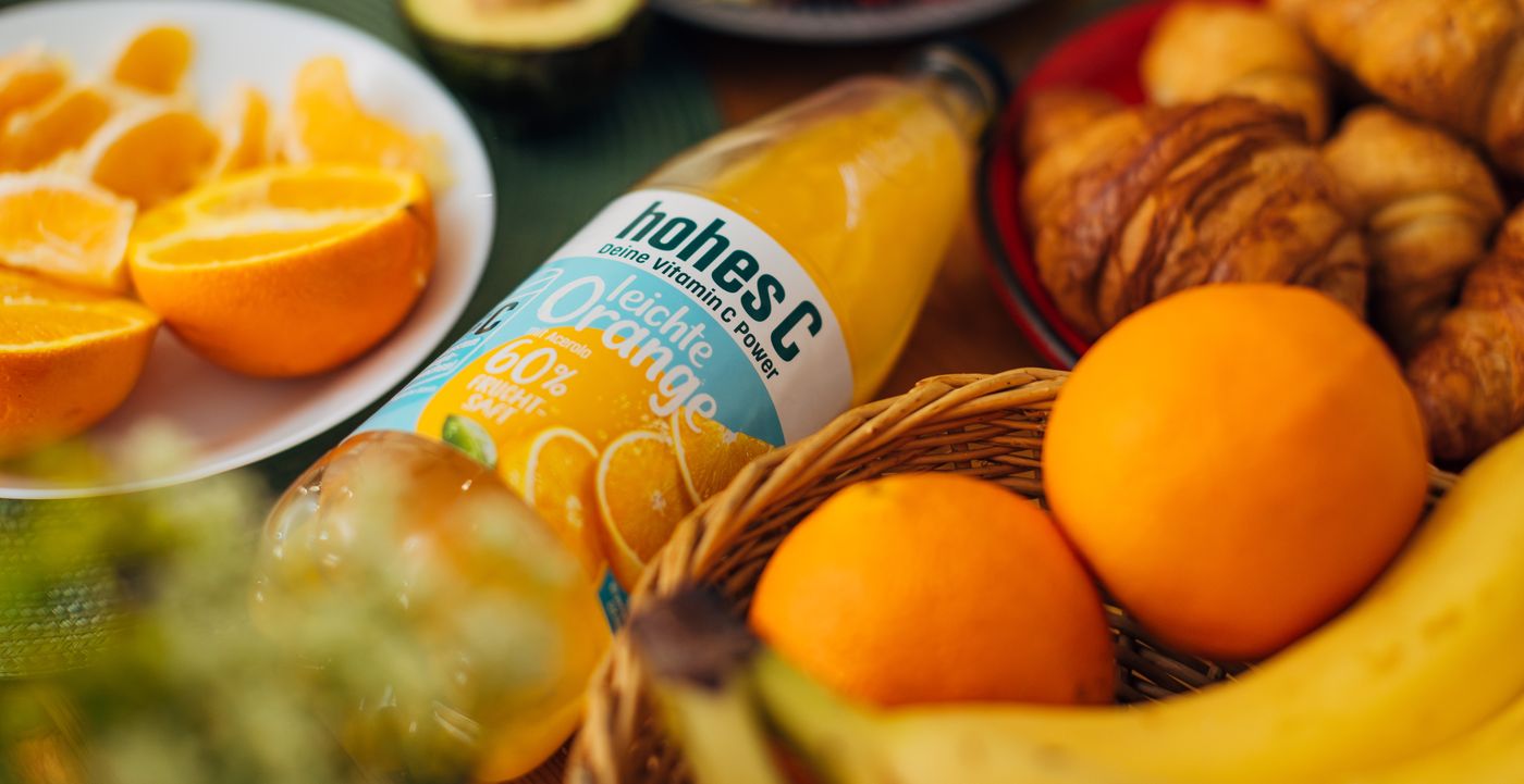 Bottle of hohes C Orange juice, croissants, whole and sliced oranges, and a banana are arranged on a table.