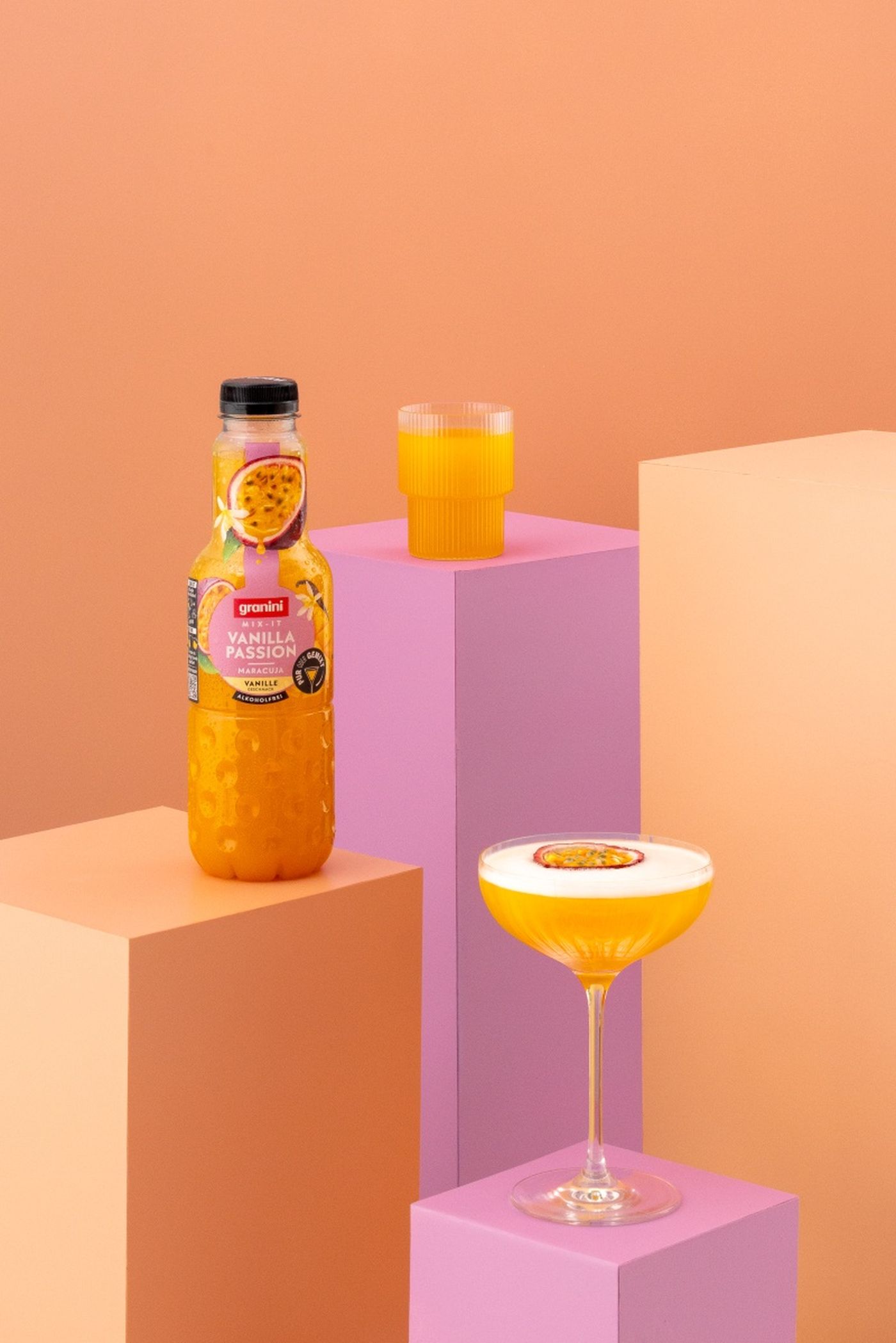 A bottle of vanilla passion fruit juice, a glass, and a cocktail glass on pastel pedestals against a peach background.