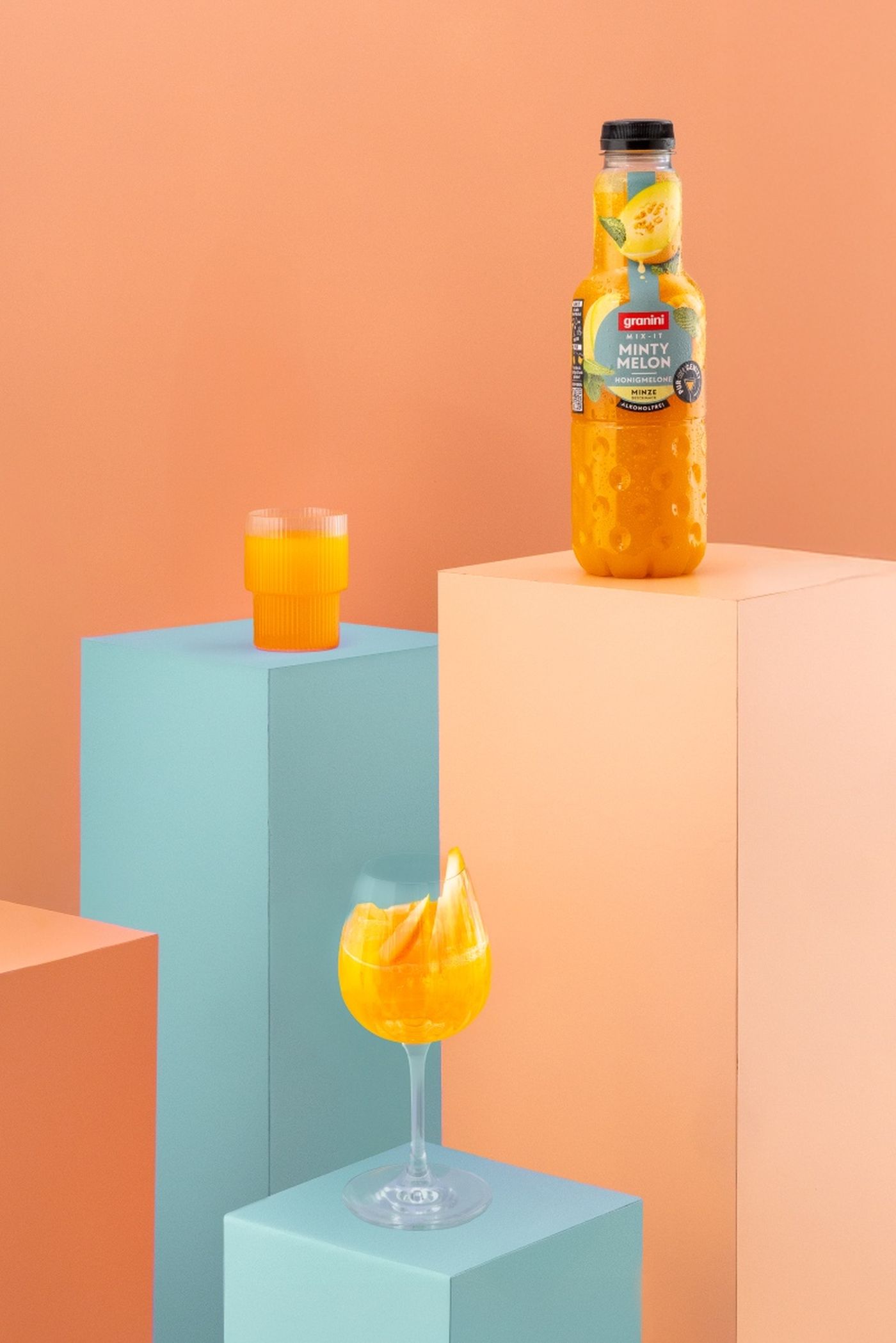 A bottle of orange juice with mint on a peach pedestal, surrounded by a glass and cup filled with orange liquid, set against colorful blocks.
