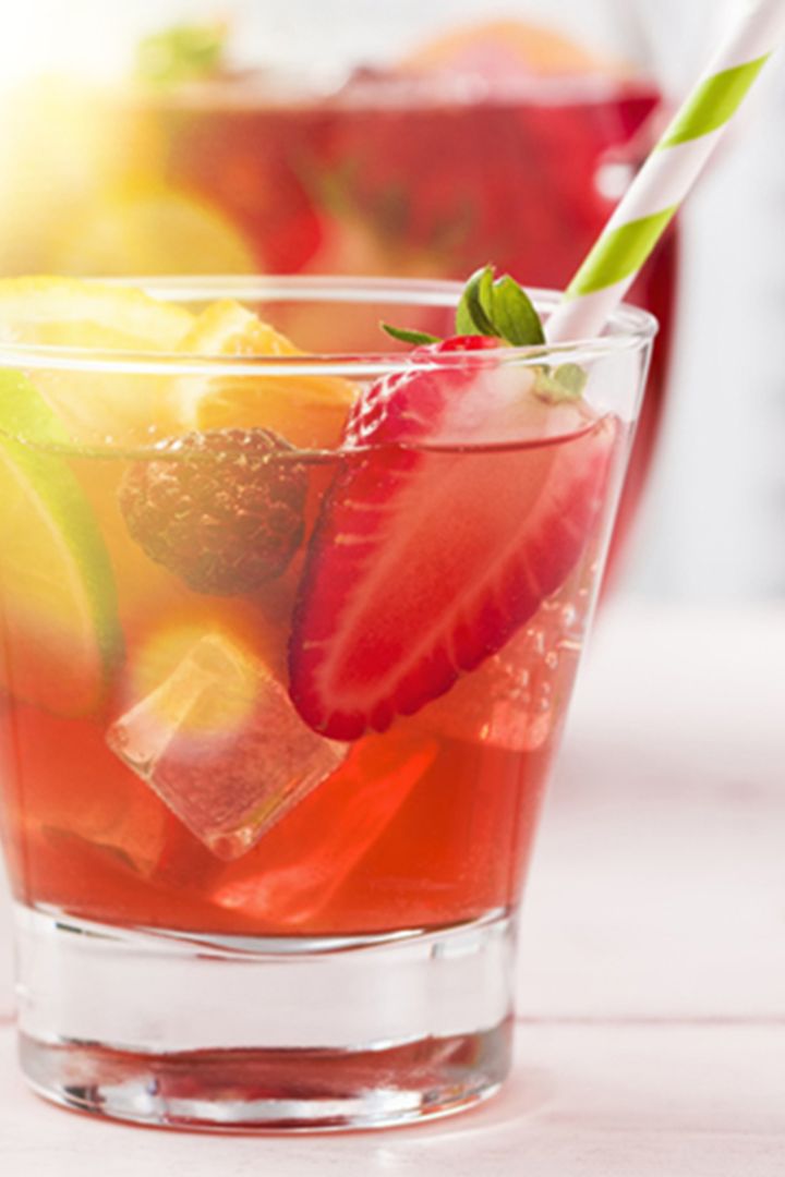 A glass of fruit punch with ice, garnished with strawberry slices, lime, and a striped straw, in soft natural light.