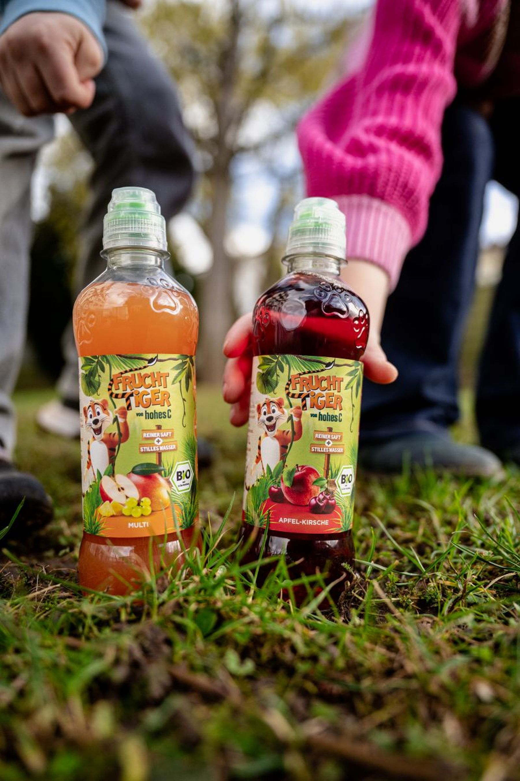 Two Frucht Tiger juice bottles, one Multi and one Apple-Cherry flavor, placed on grass with hands reaching for them.