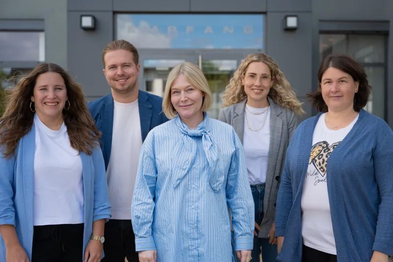 Five people smiling, standing in front of a building entrance. They are casually dressed and appear to be part of a professional group.