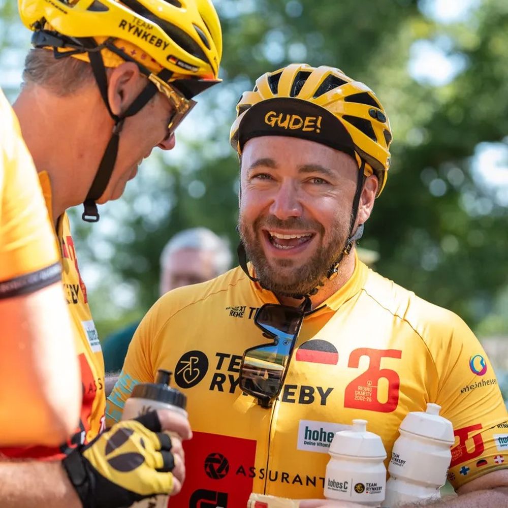 Two cyclists in yellow jerseys and helmets chat and smile outdoors, holding water bottles, with greenery in the background.