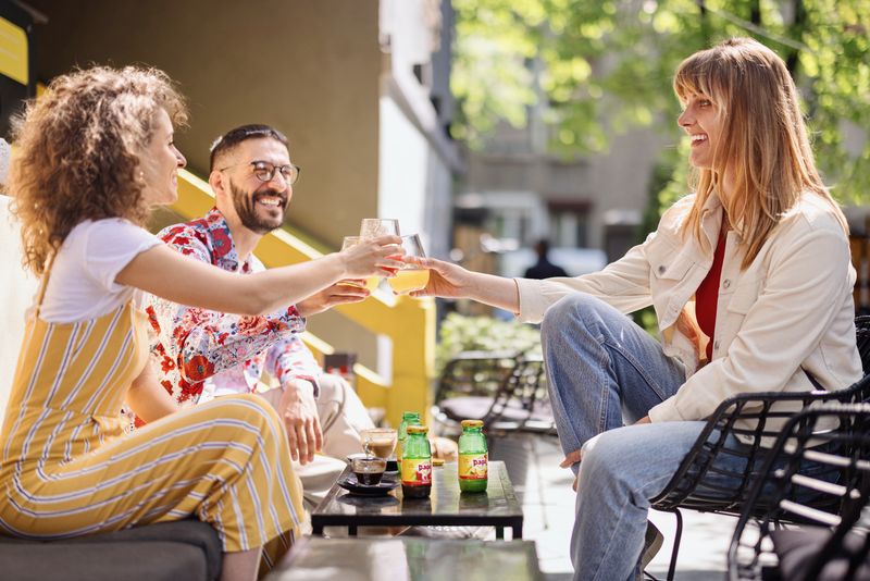 3 Personen sitzen auf Gartenmöbeln. In der Mitte steht ein Tisch, auf dem 3 Pago Flaschen und Kaffee steht. Die 3 Leute stoßen mit Gläsern an, die mit Pago Fruchtsäften gefüllt sind. Im Hintergrund ist ein Baum.