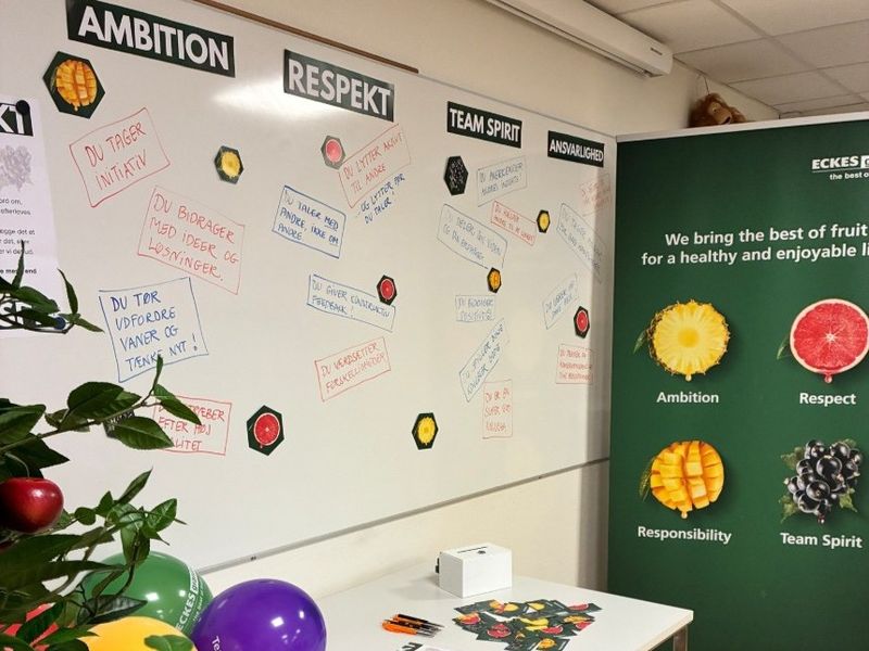 A whiteboard with sections labeled "Ambition," "Respect," "Team Spirit," and "Responsibility" has handwritten notes. Nearby is a banner with fruit images.
