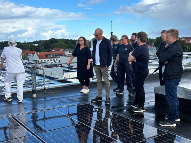 A group of people standing on a rooftop beside solar panels, with a view of a town and trees in the background under a partly cloudy sky.