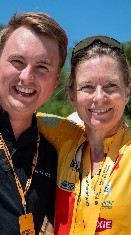 Two smiling people, one in a black shirt and the other in a yellow cycling outfit, posing outdoors on a sunny day.