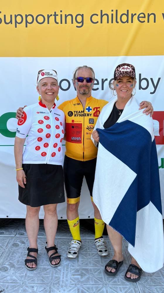 Three people smile in front of a "Supporting children" banner. Two wear Team Rynkeby cycling attire; one draped in a blue and white flag.