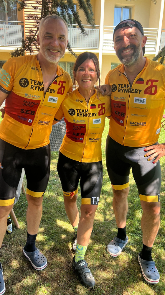 Three cyclists in matching Team Rynkeby jerseys pose and smile, standing on grass, with a building in the background.