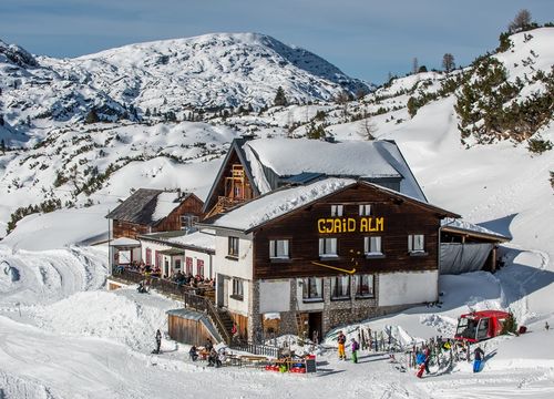 Skifahrer und Gäste vor der Gjaid Alm im Schnee, umgeben von verschneiten Bergen und Wintersonne.