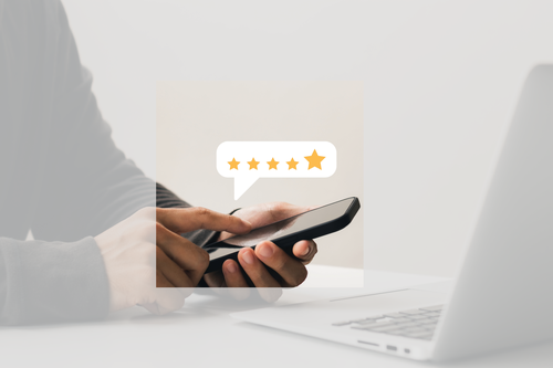 Person using a smartphone with a five-star rating bubble above it, next to an open laptop on a white desk.
