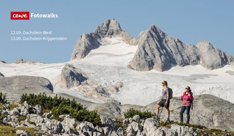 Zwei Wanderinnen auf einem felsigen Pfad mit Blick auf den beeindruckenden Dachstein-Gletscher und hohe Berggipfel. Cewe Fotowalks.